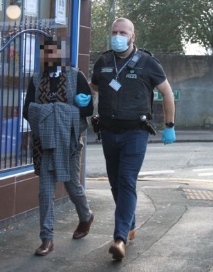 Police officer wearing mask escorts suspect past building