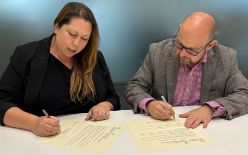 Two people sitting at a desk signing a document left woman with long dark hair and to the right man with glasses