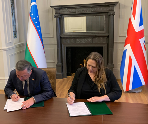 Two people sitting at a wooden desk in front of a fireplace and the Uzbekistan and UK flags signing a document. On the left Mr Musayev, Director of the Migration Agency under the Cabinet of Ministers of Uzbekistan and on the right Elysia McCaffrey CEO of the GLAA