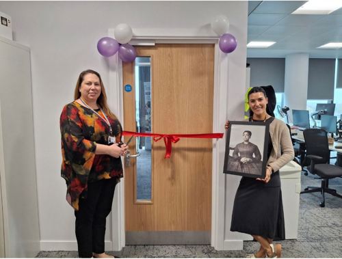 CEO Elysia McCaffrey holding a pair of scissors in front of an office door with red ribbon alongside Director Sam Dowsing holding a framed photo of Mary Prince